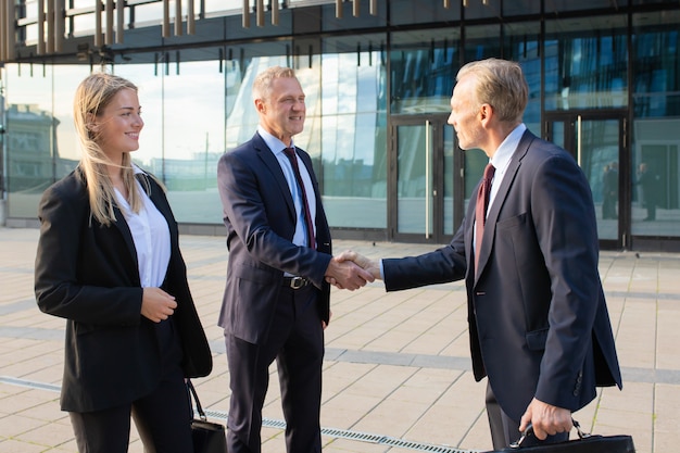 Pozytywni partnerzy biznesowi spotykają się w biurowcu, ściskają sobie ręce. Widok z boku, średni strzał. Koncepcja komunikacji korporacyjnej lub uścisk dłoni