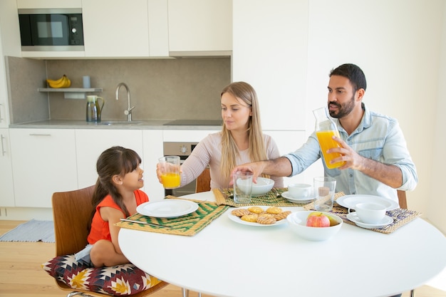 Pozytywni rodzice i córka siedzą przy stole z daniem, owocami i ciasteczkami, wlewając sok pomarańczowy.