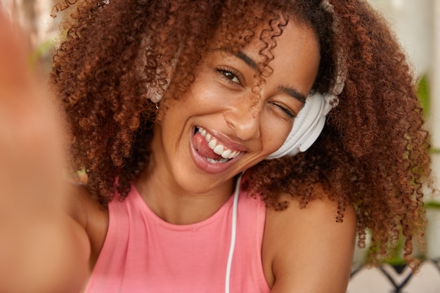 Pozytywnie kręcona Afroamerykańska dziewczyna mruga oczami, pokazuje język, jest w dobrym nastroju, słyszy melodię w słuchawkach, wyciąga rękę i robi selfie portret nierozpoznawalnym urządzeniem