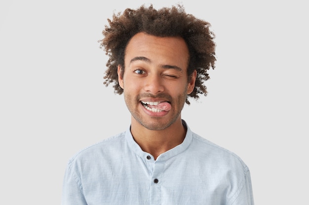 Pozytywny, atrakcyjny Afroamerykanin z pozytywnym wyrazem twarzy, pokazuje język, ma wesoły wyraz, stoi pod białą ścianą, ma chrupiące włosy