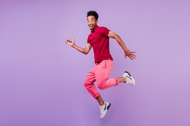 Pozytywny beztroski afrykański mężczyzna w butach sportowych, taniec. Przystojny zadowolony facet w różowe spodnie skacze z uśmiechem.