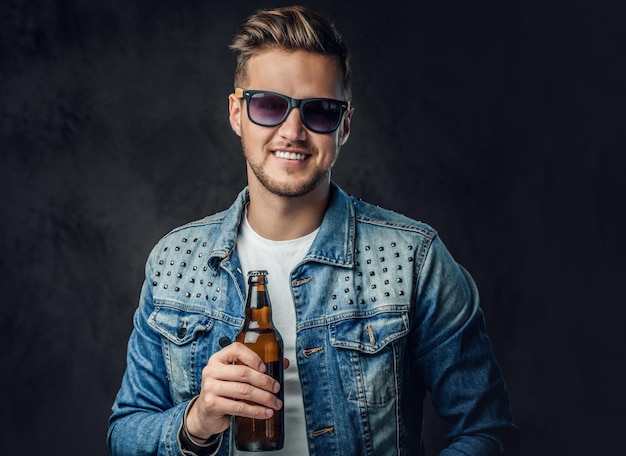 Pozytywny blond mężczyzna ubrany w dżinsową kurtkę i okulary przeciwsłoneczne trzyma butelkę z piwem rzemieślniczym.