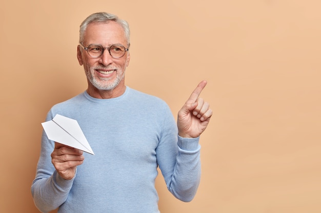Pozytywny brodaty siwowłosy starszy mężczyzna myśli o wyjeździe za granicę trzyma samolot z papieru czerpanego wskazuje w prawym górnym rogu nosi zwykły sweter odizolowany na brązowej ścianie pokazuje miejsce na kopię