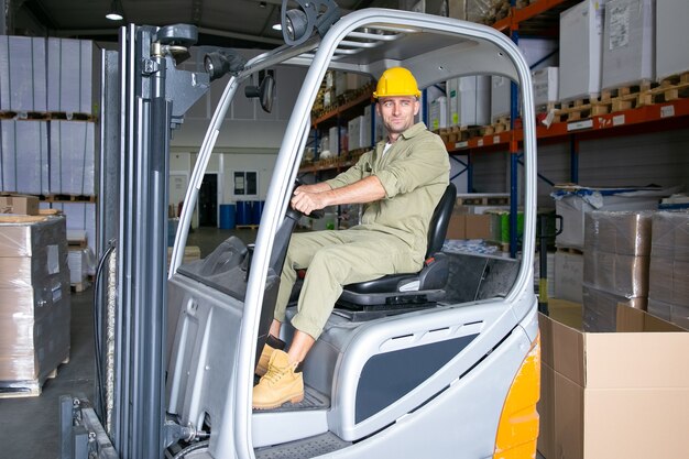 Pozytywny męski pracownik logistyczny w kask jazdy wózkiem widłowym w magazynie, uśmiechając się, odwracając wzrok