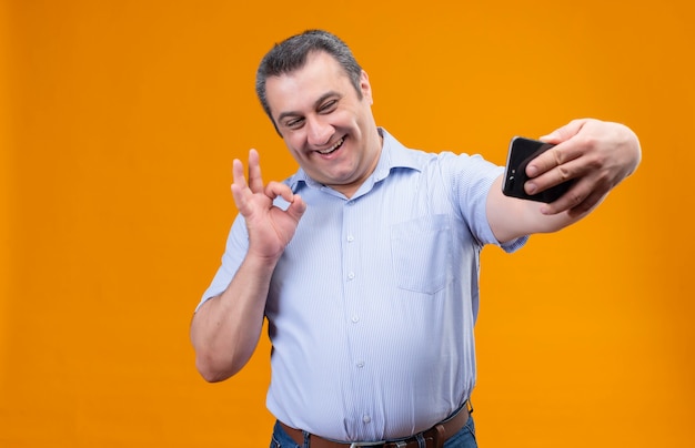 Pozytywny mężczyzna w średnim wieku w niebieskiej koszuli w pionowe paski, śmiejąc się i biorąc selfie na smartfonie na pomarańczowym tle