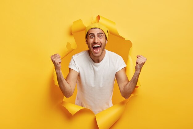 Pozytywny mężczyzna wiwatuje z zaciśniętymi pięściami, świętuje zwycięstwo, nosi zwykłą białą koszulkę i żółtą czapkę