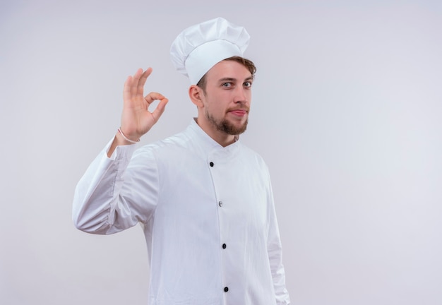 Pozytywny młody brodaty szef kuchni w białym mundurze pokazuje gest ok na białej ścianie