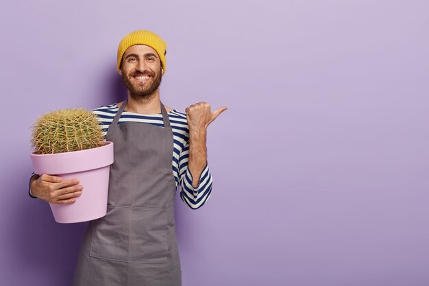 Pozytywny ogrodnik wskazuje kciukiem, pokazuje puste miejsce na reklamę, trzyma doniczkę z tłem kaktusa