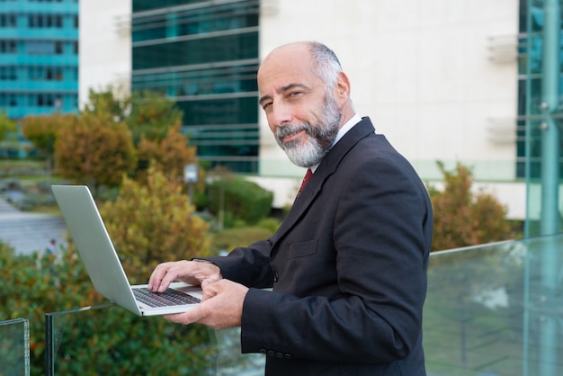 Pozytywu dojrzały biznesmen używa laptop blisko budynku biurowego