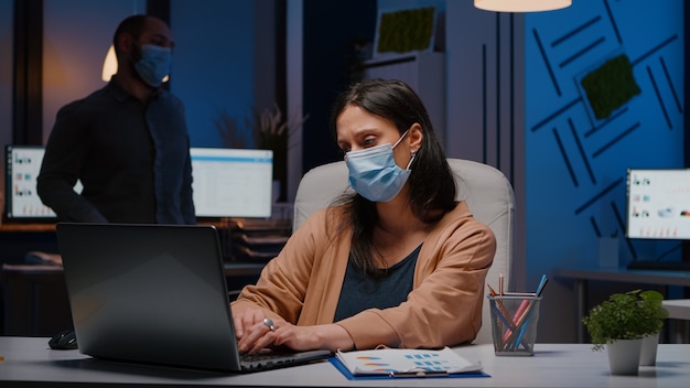 Pracoholik bizneswoman z maską na twarz przeciwko covid19 pracujący w startupowym biurze analizującym strategię ekonomiczną późno w nocy