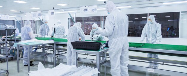 Pracownicy fabryki w białych garniturach laboratoryjnych i maskach na twarz, produkujący telewizory na zielonej linii montażowej z nowoczesnym sprzętem