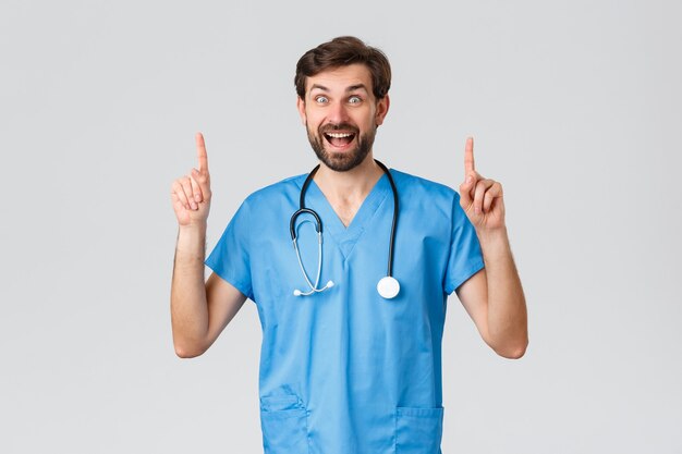 Pracownicy opieki zdrowotnej, koncepcja wybuchu pandemii i koronawirusa. Entuzjastycznie uśmiechnięty brodaty lekarz w niebieskim fartuchu, ze stetoskopem, wskazującym palcem w górę, wyświetlającym reklamę na górze, wyglądającym na zdumionego i szczęśliwego.
