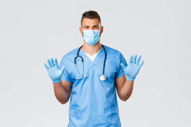 Pracownicy służby zdrowia, covid-19, koronawirus i koncepcja zapobiegania wirusowi. Poważnie wyglądający latynoski lekarz, pielęgniarz w fartuchu, maska medyczna, pokazując rękawiczki na rękach, poproś o założenie sprzętu ochronnego