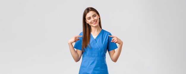Pracownicy służby zdrowia zapobiegają koncepcji wirusów i leków Wesoła uśmiechnięta ładna lekarka pielęgniarka w niebieskich fartuchach wskazująca na siebie gotowa pomóc pacjentom ratować życie przed koronawirusem