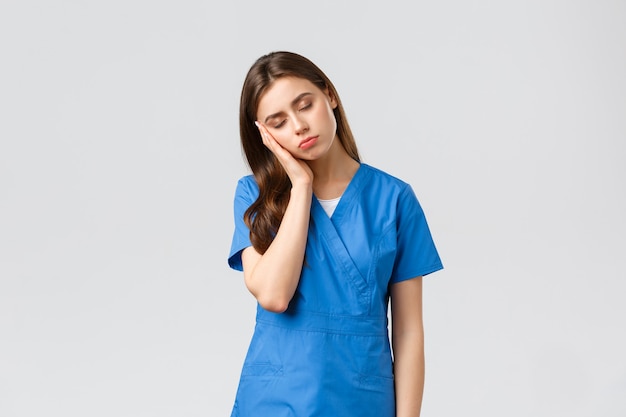 Pracownicy służby zdrowia, zapobieganie koncepcji wirusa i medycyny. Zmęczona śpiąca lekarka, młoda pielęgniarka w niebieskim fartuchu, opiera się na dłoni i zamyka oczy, wyczerpana po nocnej zmianie w klinice w walce z koronawirusem