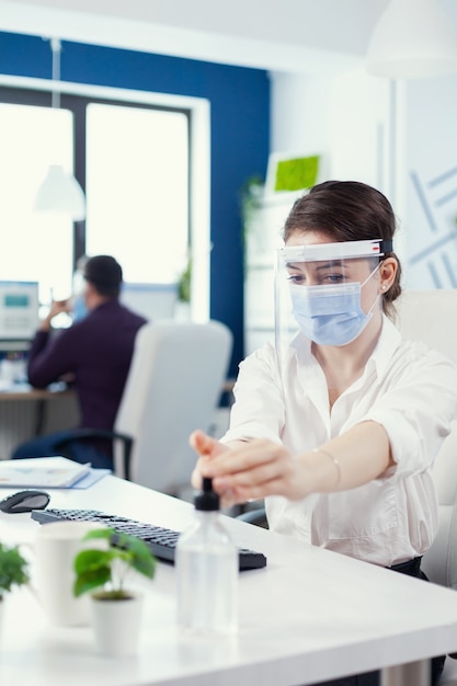 Pracownik biurowy przestrzegający środków ostrożności podczas globalnej pandemii z koronawirusem stosującym środek dezynfekujący. Businesswoman w nowym normalnym miejscu pracy dezynfekcji, podczas gdy koledzy pracujący w tle.
