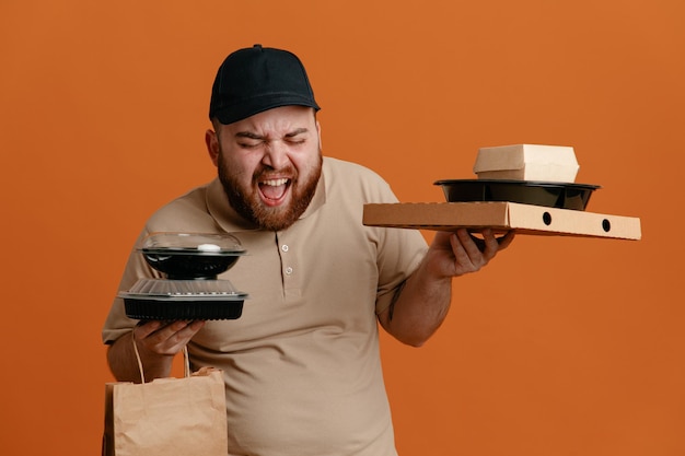 Bezpłatne zdjęcie pracownik dostawy w czarnej czapce i pustym mundurze tshirt, trzymający pojemniki na żywność z papierową torbą, wrzeszczącą i krzyczącą zły i szalony szalony stojący na pomarańczowym tle