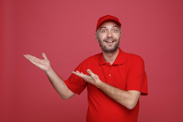 Pracownik dostawy w czerwonej czapce puste tshirt mundur patrząc na kamery szczęśliwy i pozytywny uśmiechający się radośnie prezentując ramionami rąk stojąc na czerwonym tle