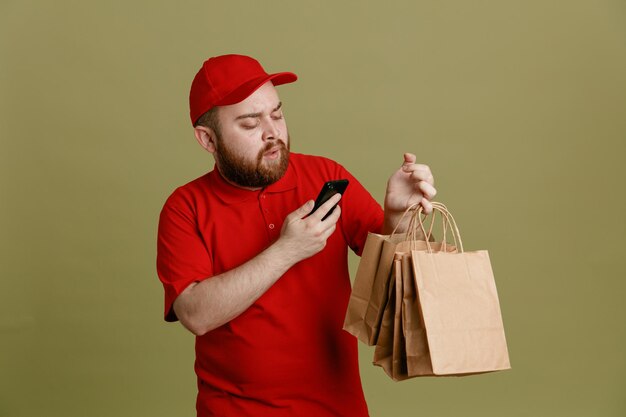 Pracownik dostawy w czerwonej czapce puste tshirt mundurze trzymając papierowe torby trzymając smartfon patrząc na ekran, będąc pewny siebie stojąc na zielonym tle