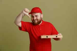 Bezpłatne zdjęcie pracownik dostawy w czerwonej czapce puste tshirt mundurze trzymając pudełko po pizzy patrząc na kamery szczęśliwy i pozytywny uśmiechający się radośnie pokazując kciuk do góry stojący na zielonym tle