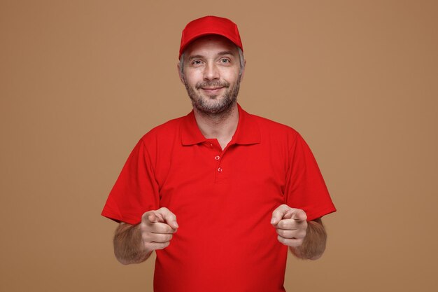 Pracownik dostawy w czerwonej czapce pusty tshirt mundurze patrząc na kamery szczęśliwy i pozytywny uśmiechający się wskazując palcami wskazującymi obiema rękami w aparacie stojącym na brązowym tle