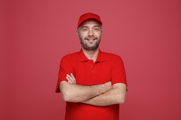 Pracownik dostawy w czerwonej czapce pusty tshirt mundurze patrząc na kamery z rękami skrzyżowanymi na piersi uśmiechnięty radośnie wyrażenie stojąc na czerwonym tle