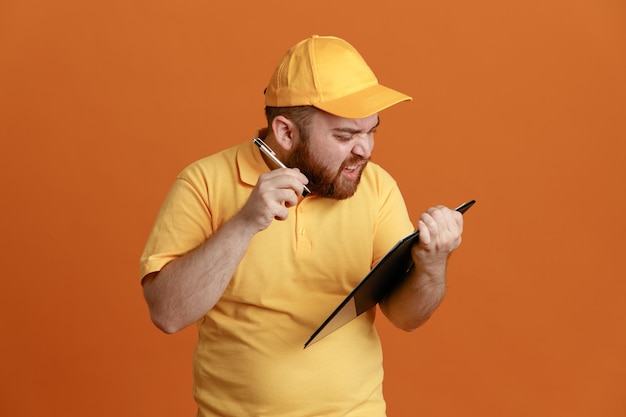 Bezpłatne zdjęcie pracownik dostawy w mundurze pustej koszulki z żółtą czapką, trzymając schowek i długopis, patrząc zdezorientowany i niezadowolony, stojąc na pomarańczowym tle