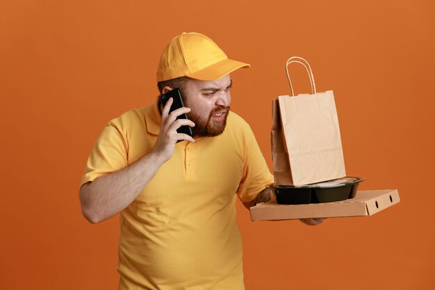 Pracownik dostawy w mundurze żółta czapka pusta koszulka, trzymając pojemnik na jedzenie i pudełko na pizzę z papierową torbą, rozmawiając na telefonie komórkowym, patrząc niezadowolony, stojąc na pomarańczowym tle