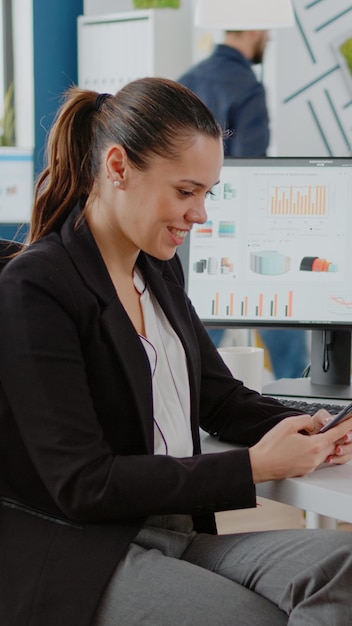 Pracownik firmy patrzący na ekran smartfona podczas pracy nad planowaniem biznesowym z komputerem i wykresami w biurze firmy. Kobieta za pomocą telefonu komórkowego do pomysłów burzy mózgów