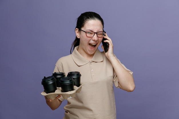 Pracownik kobieta dostawy w pustym mundurze koszulki polo w okularach, trzymając filiżanki kawy, krzycząc z zirytowanym wyrazem twarzy podczas rozmowy na telefonie komórkowym stojącym na niebieskim tle