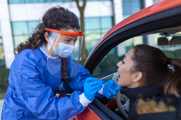 Pracownik medyczny przeprowadzający kontrolę COVID19 przez automobilizm pobierający próbkę wymazu z nosa od pacjentki przez okno samochodu Diagnostyka PCR pod kątem obecności koronawirusa w zestawie testowym zawierającym PPE