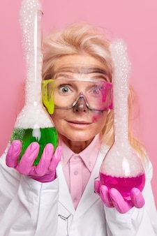 Profesjonalna chemia kobieca trzyma dwie kolby z kolorowym płynem pokazuje eksperyment chemiczny w laboratorium nosi okulary ochronne białą szatę i gumowe rękawiczki brudne po niespodziewanym wybuchu