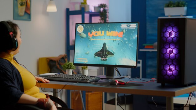 Profesjonalna grająca w e-sporty kobieta grająca na potężnej komputerowej grze wideo RGB, świętująca zwycięstwo. Pro cyber-streaming podnoszący ręce wygrywając turniej, e-sportowe mistrzostwa online od studia gier