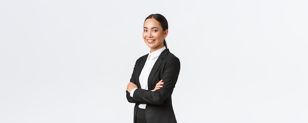 Profesjonalna młoda azjatycka kobieta-przedsiębiorca agent real eastate w garniturze, skrzyżowane ramiona i patrząca pewnie w kamerę, odnosząca sukcesy bizneswoman, prowadząca biznes, prowadząca zespół, rozpoczynające spotkanie