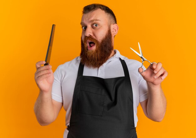 Profesjonalny brodaty fryzjer w fartuchu trzymając nożyczki i szczotkę do włosów zaskoczony stojąc nad pomarańczową ścianą