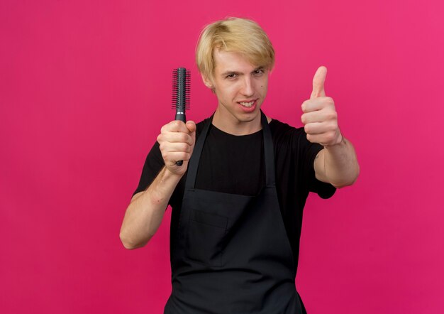 Profesjonalny fryzjer mężczyzna w fartuch trzymając szczotkę do włosów, uśmiechając się, pokazując kciuki do góry