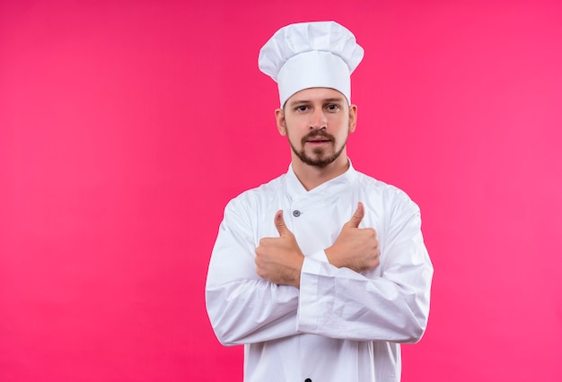 Profesjonalny kucharz mężczyzna w białym mundurze i kapelusz kucharz skrzyżowanie rąk pokazując kciuki do góry patrząc pewnie stojąc na różowym tle