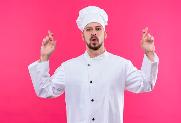 Profesjonalny kucharz mężczyzna w białym mundurze i kapeluszu kucharskim sprawia, że pożądane życzenie trzyma kciuki stojąc na różowym tle