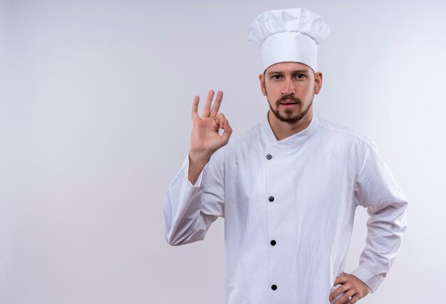 Profesjonalny kucharz mężczyzna w białym mundurze i kucharz kapelusz wskazujący znak, patrząc pewnie stojąc na białym tle