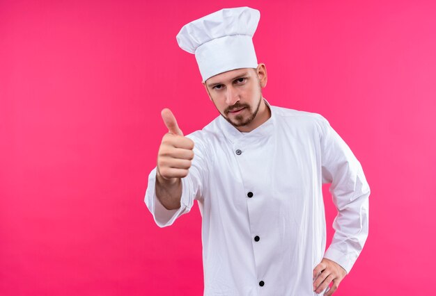 Profesjonalny mężczyzna kucharz w białym mundurze i kapelusz kucharz patrząc na kamery pokazując kciuki stojąc na różowym tle
