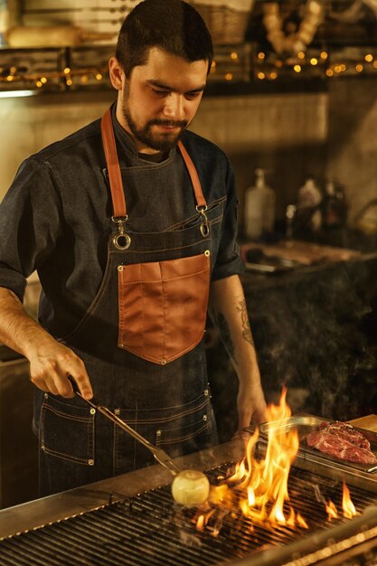 Profesjonalny szef kuchni gotuje cebulowe warzywa i mięso na grillu z ogniem i dymem piękny mężczyzna skoncentrowany na przygotowywaniu żywności w nowoczesnej kuchni restauracyjnej