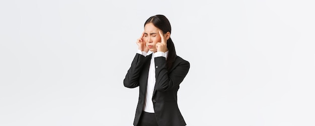 Profil azjatyckiej bizneswoman w czarnym garniturze z migreną dotykającą świątyń i grymasem z bolesnego uczucia w głowie Kobieta kierownik biura mająca ból głowy potrzebuje środków przeciwbólowych na białym tle