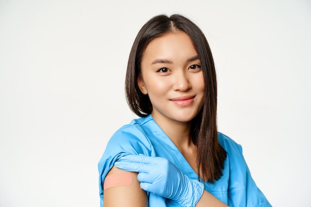 Program szczepień i koncepcja Covid-19. Portret azjatyckiego pracownika służby zdrowia w medycznej szacie, pokazujący zaszczepione ramię i uśmiechnięty, stojący na białym tle