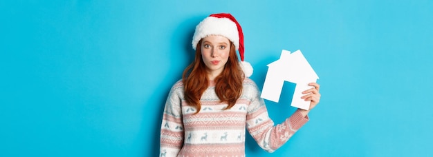 Bezpłatne zdjęcie promocje wakacyjne i koncepcja nieruchomości śliczna rudowłosa kobieta w santa hat i sweter przedstawiający papierowy hou