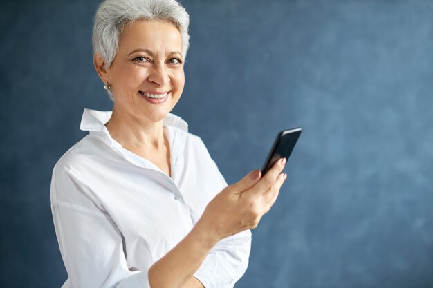przedsiębiorca kobieta w średnim wieku z krótkimi siwymi włosami trzymająca telefon komórkowy, wykonująca połączenia biznesowe, pisząca wiadomość tekstową.