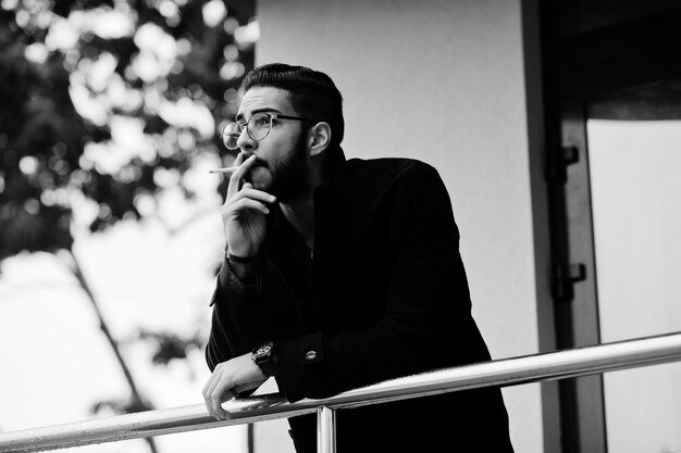 Przedsiębiorca z Bliskiego Wschodu nosi czarny płaszcz i okulary w niebieskiej koszuli przed biurowcem palącym papierosy