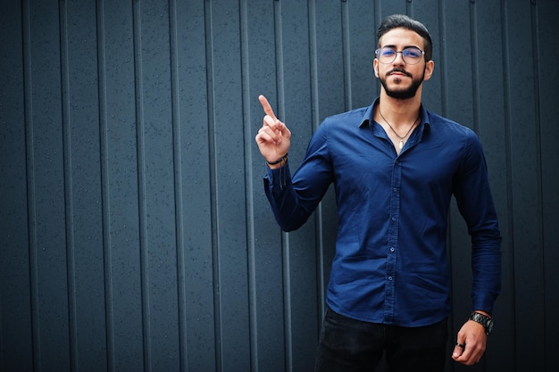 Przedsiębiorca z Bliskiego Wschodu nosi okulary w niebieskiej koszuli na tle stalowej ściany, pokazując palec w górę