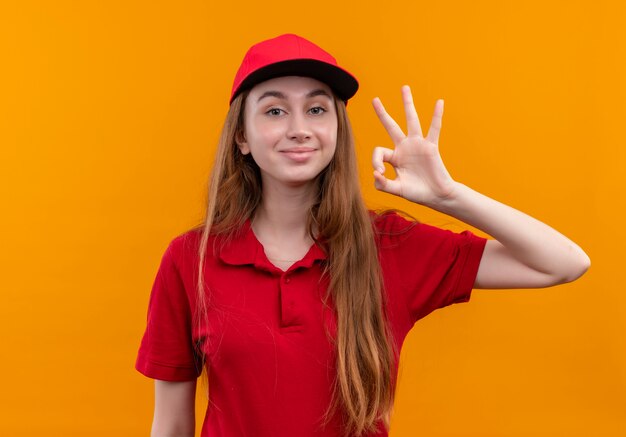 Przekonana, młoda dziewczyna dostawy w czerwonym mundurze robi ok znak na odizolowanej pomarańczowej przestrzeni