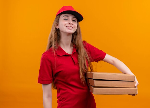 Przekonana, młoda dziewczyna dostawy w czerwonym mundurze, trzymając pakiety na odizolowanej przestrzeni pomarańczowy