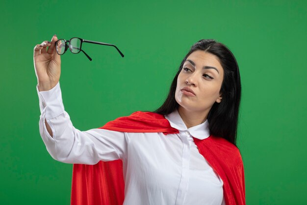 Przekonana, młoda dziewczyna superbohatera kaukaski patrząc na okulary, które trzyma w ręku na białym tle na zielonej ścianie
